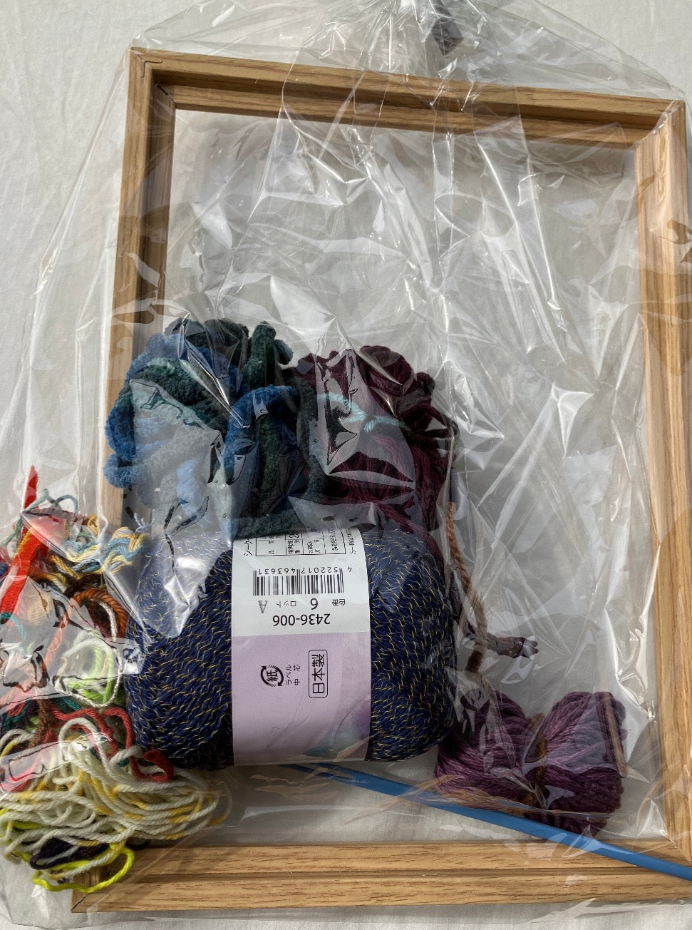 Plastic wrapped weaving kit with colorful yarn, blue plastic needle, and wooden frame